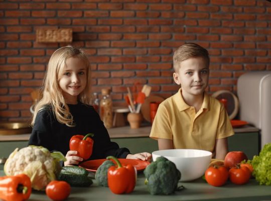 Positive little girl and boy sitting at table with various vegetables and herbs while preparing vegetarian meal in loft kitchen with brick wall