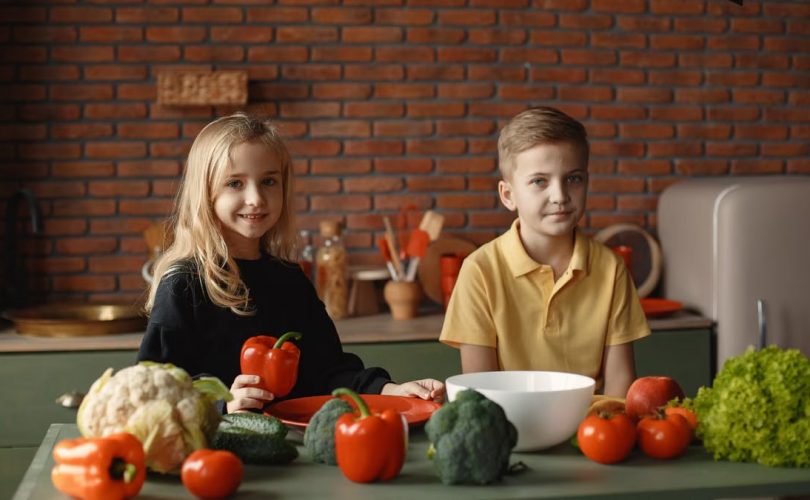 Positive little girl and boy sitting at table with various vegetables and herbs while preparing vegetarian meal in loft kitchen with brick wall